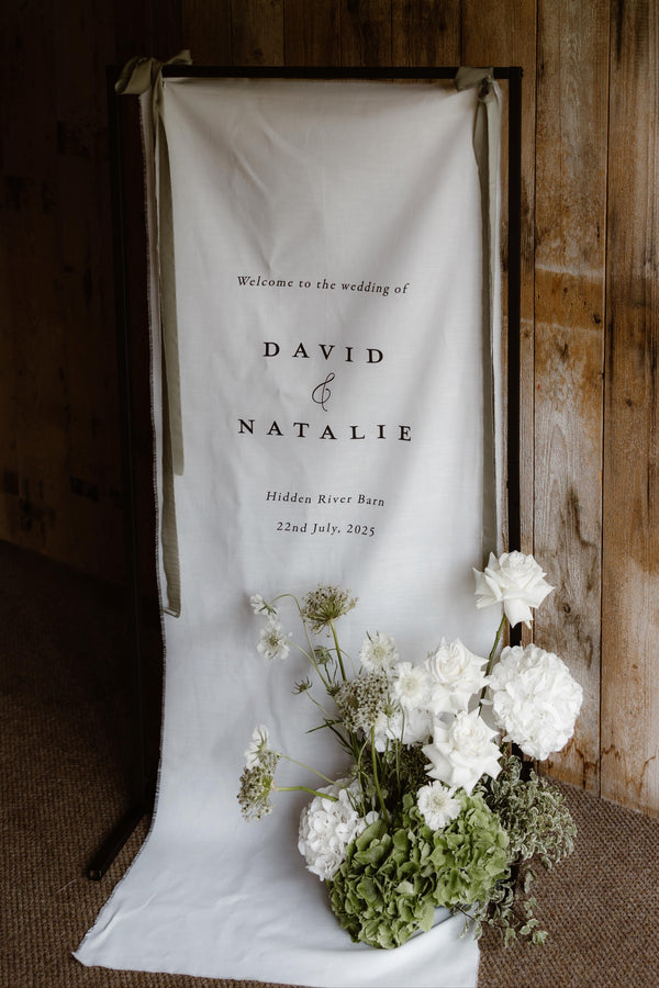 Wedding welcome sign with 'David & Natalie' and floral arrangement in front of wooden wall.